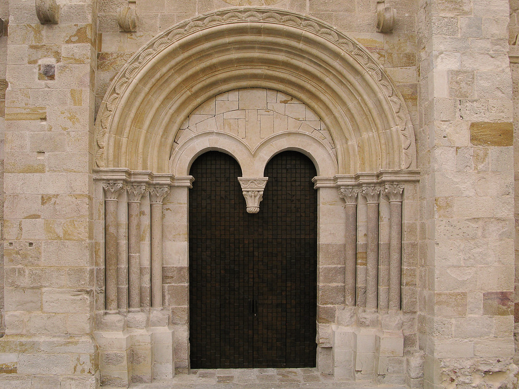 Portada románica de arquivoltas, con dos arcos gemelos sobre columnas y puerta claveteada, en un muro de sillería.